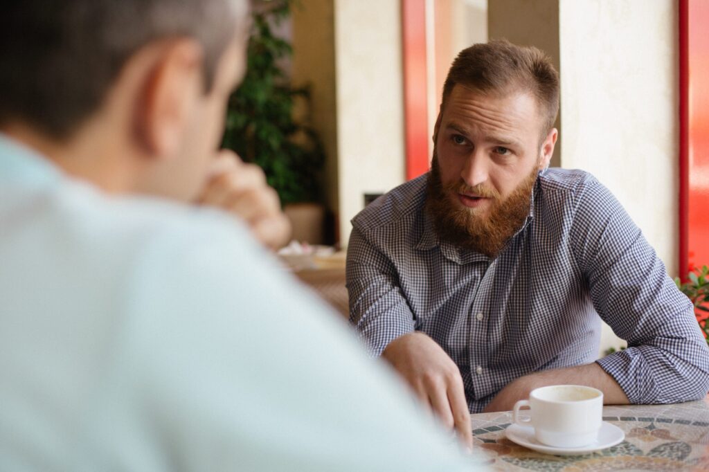young bearded man explain something to his interlocutor friend sitting indoors cafe with cup of coffee